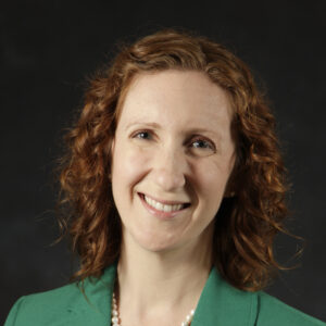 A woman with curly red hair smiles warmly, wearing a green blazer and a pearl necklace.