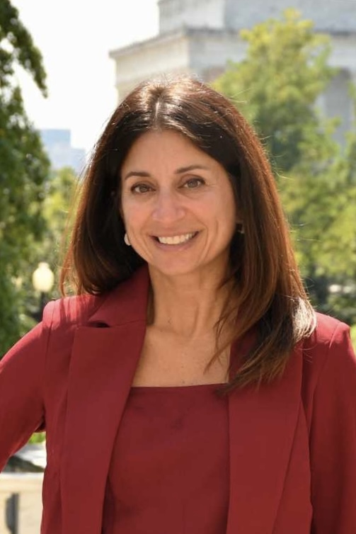 A woman in a red blazer and top smiles outside. Trees and a blurred building are in the background.
