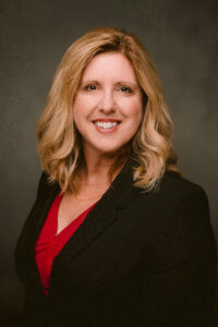 Smiling woman with shoulder-length blonde hair, wearing a black blazer over a red top, against a neutral background.