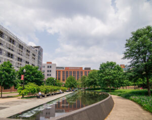 A curving pathway and reflective water feature lead through a lush green park flanked by a modern building, with a partly cloudy sky overhead.