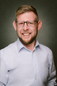 Person with short hair, glasses, and a beard smiling in a portrait against a dark background