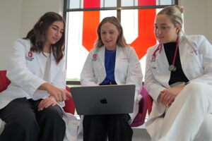Three women in white lab coats engage with a laptop, sitting together on red chairs. They appear focused, suggesting a collaborative medical setting.