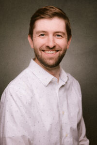 A smiling man with a beard in a white shirt with small patterns stands against a gray background, exuding warmth and friendliness.