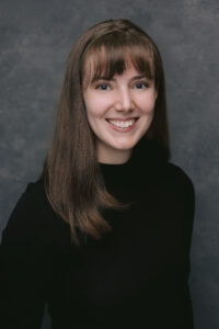 Professional headshot of a person with shoulder-length brown hair wearing a black top.