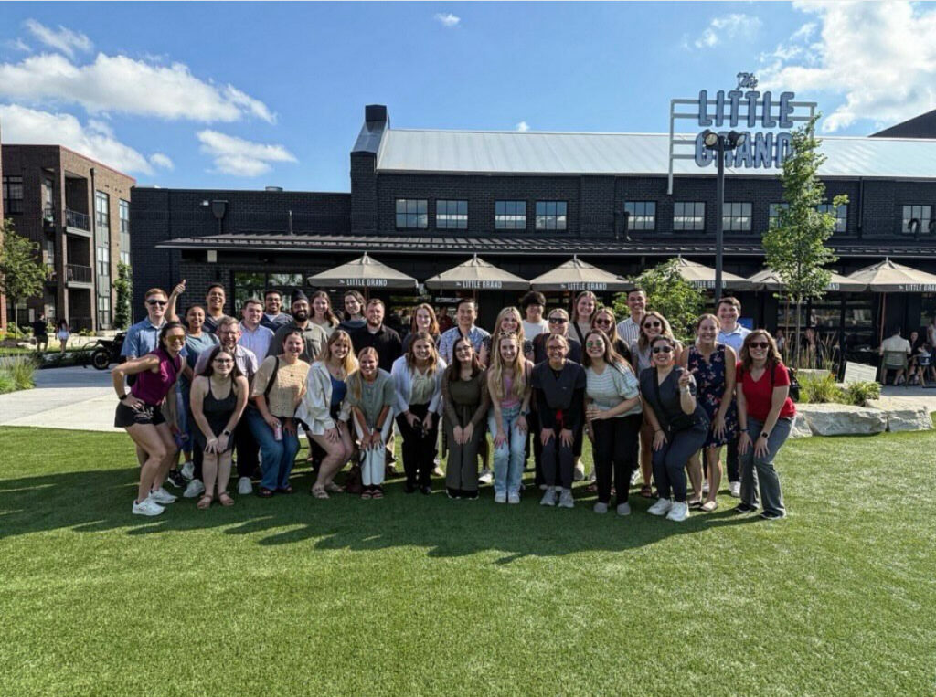 A large group of people smiles while posing on a grassy area in front of a building with "The Little Grand" sign