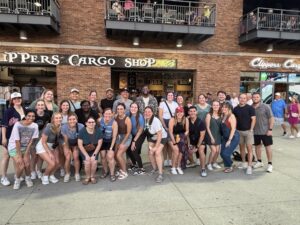 A diverse group of about 30 people smiling and posing in front of a brick building labeled "Clippers Cargo Shop." The atmosphere is lively and cheerful.