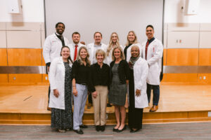 A group of ten people stand smiling on a stage. Several wear white coats.