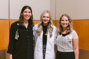 Three women stand smiling. The center woman wears a white coat; the others wear business attire.