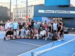 A large group of people smile and pose on a blue pickleball court at Gravity Experience Park. They appear casual and happy, with a dog joining them.