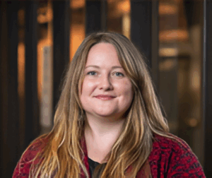 A person with long hair smiles warmly, wearing a red patterned jacket.