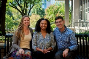 Three people sit on a bench outdoors, smiling. Lush green trees and a modern building surround them.