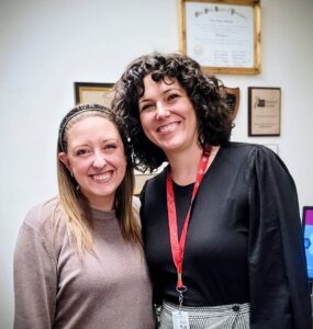 Two women smiling warmly in an office setting, surrounded by awards and certificates