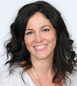 A smiling woman with wavy, dark hair wears a light top and a gold necklace