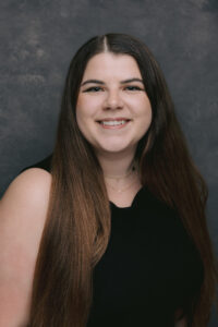 Professional headshot of a person with long dark hair wearing a black sleeveless top.