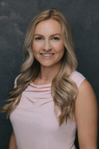 Professional headshot of a person with long wavy hair wearing a light pink sleeveless top.