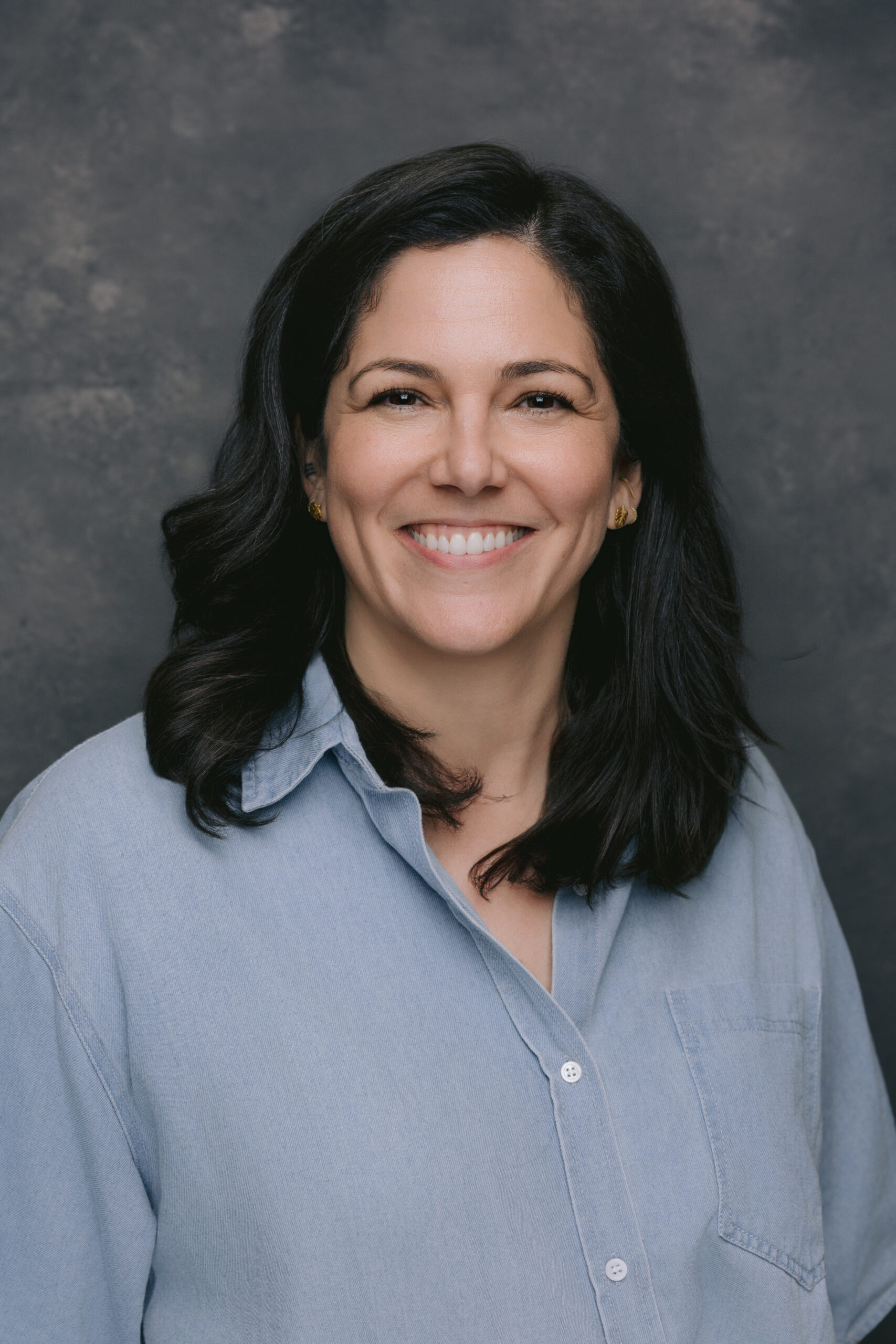 Professional headshot of a person with shoulder-length dark hair wearing a light blue button-down shirt.