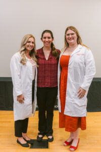 Three people standing together on a stage, with two wearing white pharmacy lab coats.