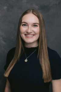 Professional headshot of a person with long straight hair wearing a black top and pendant necklace.