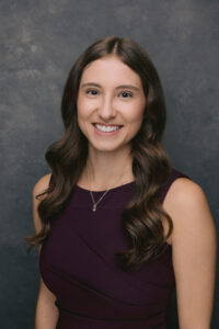 Professional headshot of a person with long curly hair wearing a sleeveless burgundy top.