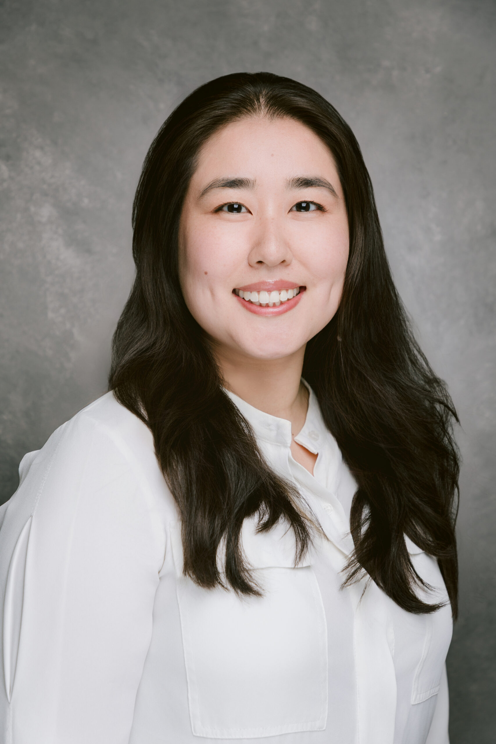 Professional headshot of a smiling person with long dark hair wearing a white blouse.