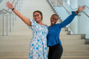 Two people stand on an indoor staircase with arms outstretched, posing for a photo.